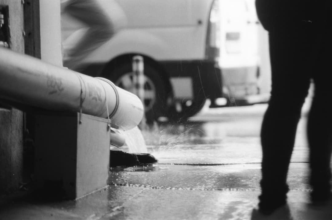 Écoulement d'eau provenant d'une canalisation avec des silhouettes floues en arrière-plan, en noir et blanc.